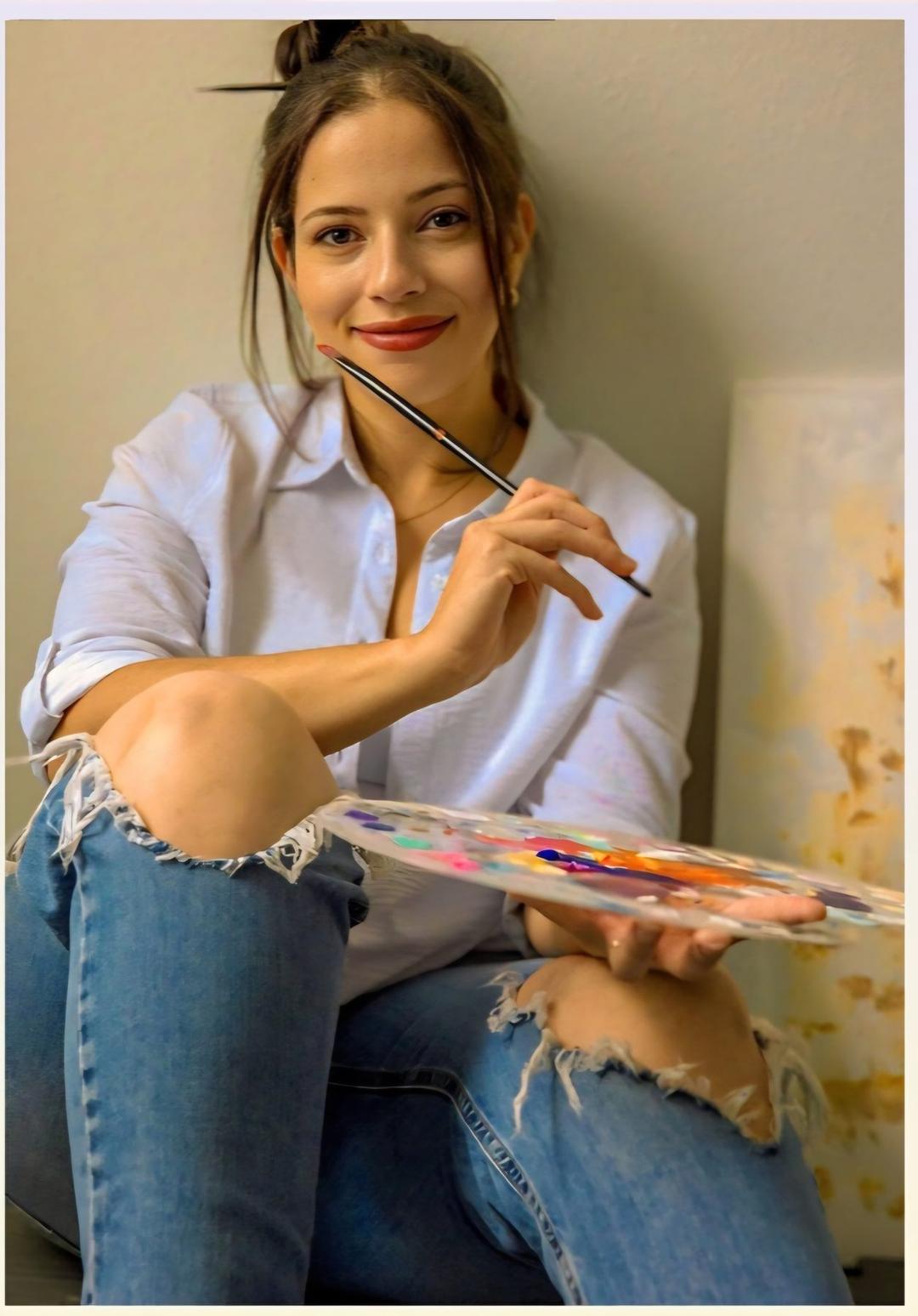 Marina Bacci smiling, holding a paintbrush and a palette of mixed colors in her studio.